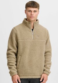 Beiger Fleece-Pullover mit hohem Kragen, halbmirtelbarem Reißverschluss und strukturiertem Material. Verfügt über seitliche Taschen und elastische Bündchen.