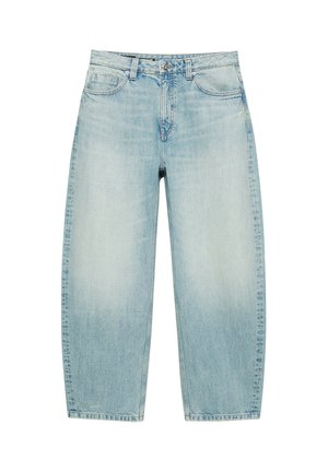 Person iført en brun stribet kortærmet skjorte over en hvid langærmet top med lyseblå jeans mod en ensfarvet baggrund.