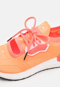 Oranžová sportovní bota s vrchní částí z prodyšné síťoviny, výrazným výřezem v designu, plochými tkaničkami a růžovými akcenty. Obsahuje bílou podrážku a gumovou spodní část pro lepší přilnavost.