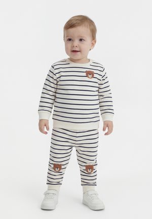 Miminko oblečené v bielom odeve s tmavomodrými vodorovnými pruhmi a hnedými nášivkami medveďa na hrudi a kolenách, stojace a pozerajúce sa doprava.