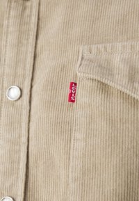 Tissu en velours côtelé beige avec des rainures verticales visibles, un bouton-pression, un rabat de poche et une petite étiquette rouge avec un texte blanc sur le côté droit.