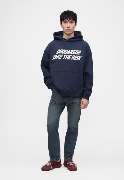 Jeune homme portant un sweat à capuche bleu marine avec le texte "DSQUARED2 TAKE THE RISK", jean bleu et baskets rouges, debout devant un fond blanc uni.