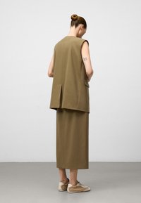 Donna con chignon, che indossa un lungo gilet senza maniche marrone e una gonna coordinata, sneakers beige, in piedi contro un muro bianco, mostrando la schiena e i tatuaggi.
