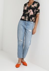 Camicetta nera a maniche corte con motivi floreali di rose rosa, abbinata a jeans leggeri azzurri strappati e scarpe arancioni senza lacci.