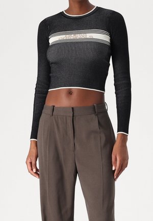 Woman wearing a black, ribbed, long-sleeve crop top with "Guess Originals" text and high-waisted brown trousers against a plain background.