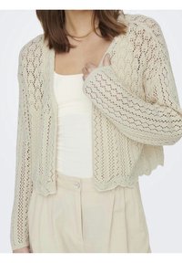 Cárdigan de punto color crema con diseño calado, manga larga, dobladillo con borde ondulado y patrón texturizado. Llevado sobre una camiseta blanca.