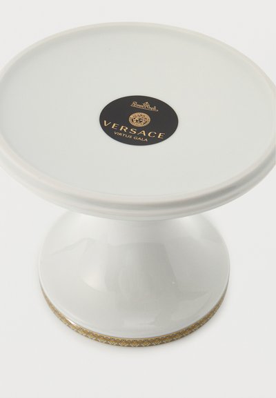 Λευκή κεραμική βάση για τούρτα με χρυσή διακοσμητική μπορντούρα και μαύρη ετικέτα με την επιγραφή "Versace Virtus Gala" στην κορυφή.
