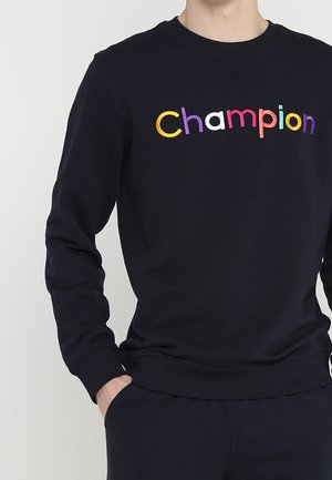 Persoon draagt een zwarte trui met "Champion" in kleurrijke letters over de borst, hand in de zak, tegen een effen achtergrond.