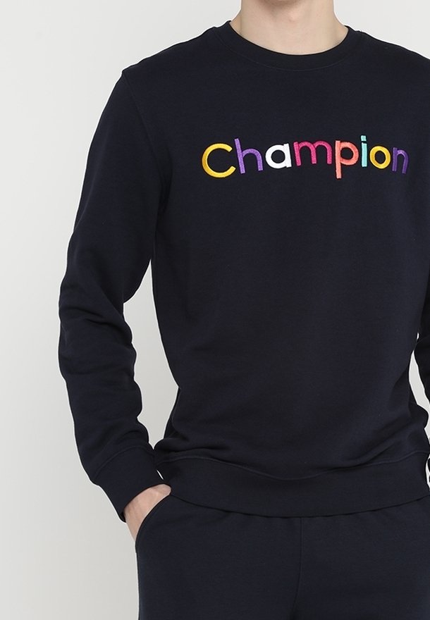 Personne portant un sweat-shirt noir avec le mot « Champion » écrit en lettres colorées sur la poitrine, main dans la poche, sur un fond uni.