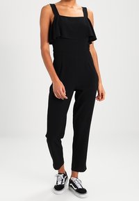 Czarny kombinezon z szerokimi ramiączkami i falbanką przy dekolcie. Dopasowana góra przechodzi w zwężane nogawki, dopełnione casualowymi czarnymi sneakerami.