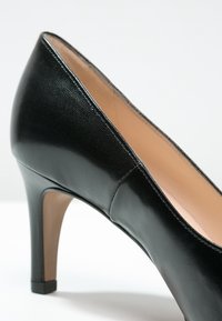 Chaussure à talon haut en cuir noir avec un bout pointu, une surface texturée et un talon ressemblant à du bois ; l'intérieur est doublé d'un beige lisse.