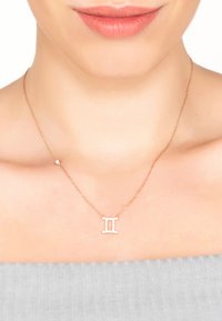 Rose gold necklace with a Gemini symbol, featuring small clear gemstones, on a delicate chain against a light gray top.