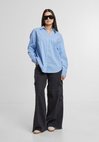 Camicia azzurra chiara, con bottoni, taglio morbido, abbinata a pantaloni cargo neri con tasche e sneakers bianche con suola marrone.