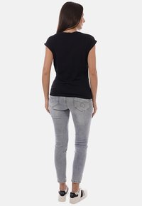 Donna con capelli lunghi e scuri, indossa una maglietta nera, jeans grigio chiaro e scarpe da ginnastica bianche, in piedi di spalle su uno sfondo neutro.