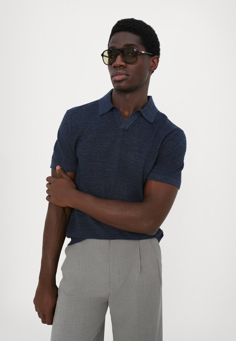 Polo a maniche corte blu con finitura tessurizzata e colletto a due bottoni, abbinato a pantaloni grigi chiari su misura. Occhiali da sole scuri completano il look.