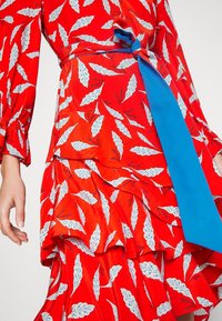 Rotes Kleid mit weißen Blättermustern, Rüschenlagen und einem blauen Band, das um die Taille gebunden ist. Lange Ärmel mit gerafften Manschetten.