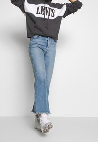 Felpa grigia con cappuccio e logo LEVI'S bianco, abbinata a jeans chiari svasati con orli sfrangiati e sneakers bianche con suola nera.
