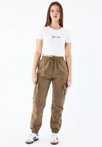 Camiseta corta blanca con texto negro, combinada con pantalones cargo oliva con bolsillos laterales; el modelo lleva zapatillas blancas y negras.