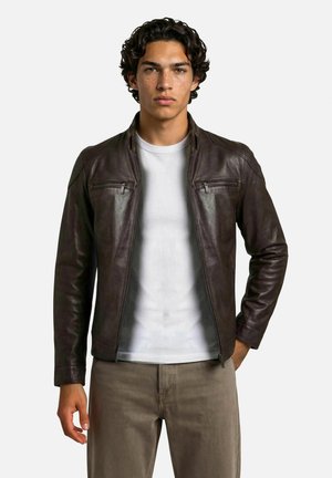 Jeune homme aux cheveux bouclés et foncés portant une veste en cuir marron foncé, un t-shirt blanc et un pantalon beige, debout devant un fond uni.