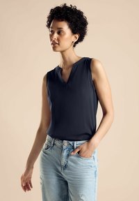 Ärmlös mörkblå blus med V-ringning, slät textur och subtila sömnadsdetaljer, kombinerad med ljusblå slitna jeans.