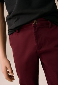 Pantalon bordeaux à texture lisse, avec une fermeture à un bouton et des passants pour ceinture. Associé à une chemise noire, offrant une coupe décontractée.