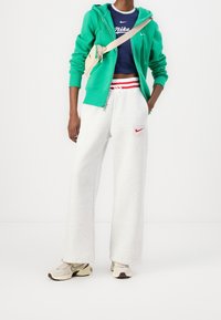 Grön zip-up hoodie, marinblå Nike crop top, grå vida sweatpants med röda detaljer och beige Nike sneakers. Synliga logodetaljer.