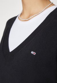 Personne portant un pull noir à col en V sur une chemise blanche avec un petit logo rouge, blanc et bleu sur la poitrine et un collier de perles.