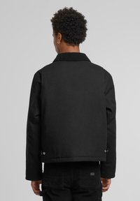 Person with short curly hair wearing a black jacket with snap buttons on the sides and black pants, viewed from the back.