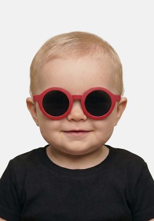 Toddler with short blonde hair wearing round red sunglasses and a black shirt, smiling against a plain light background.