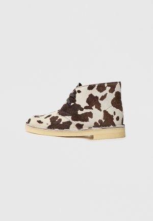 Ghete de gleznă din piele de vacă maro și crem, cu vârf rotund, design cu șireturi și talpă din cauciuc deschis la culoare. Suprafață texturată cu modele naturale.