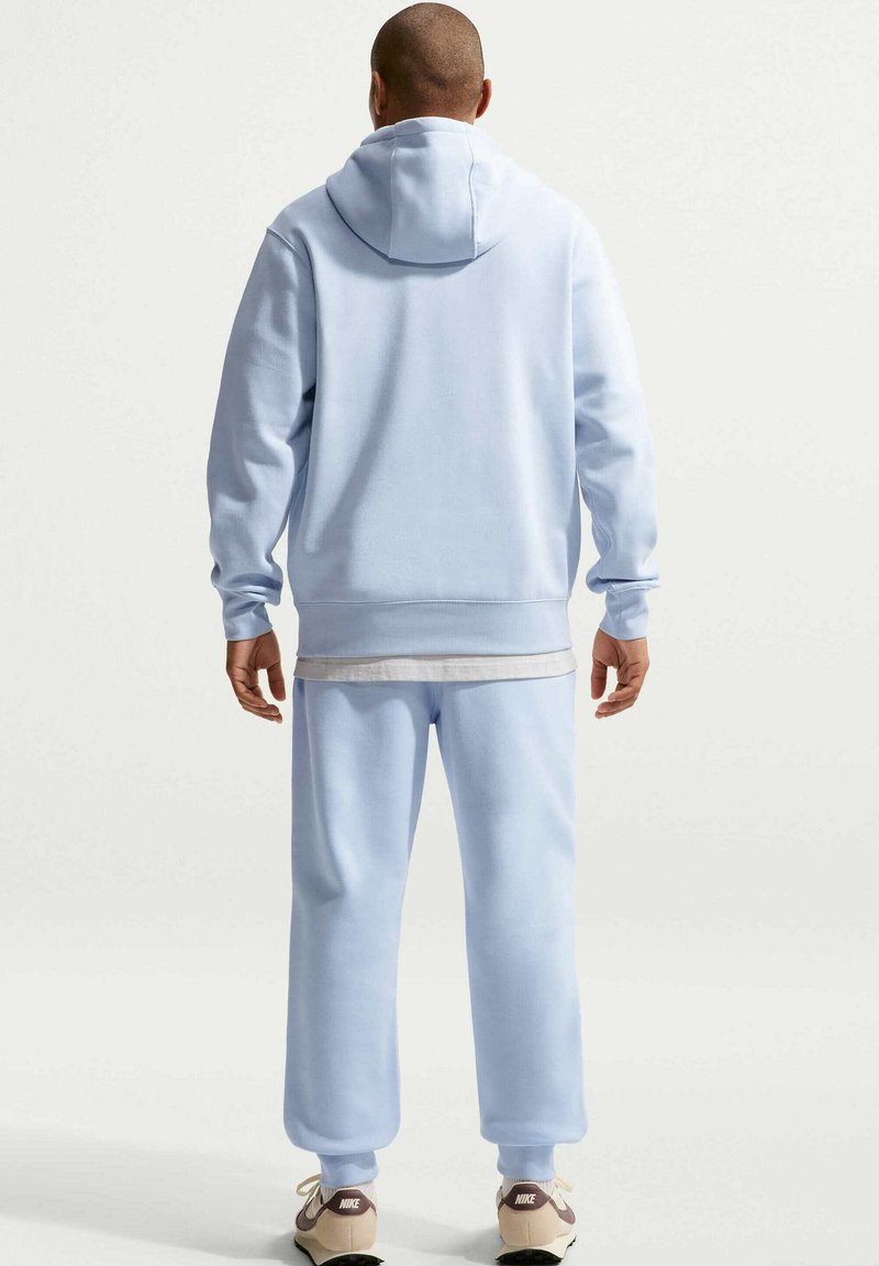 Uomo in piedi di spalle alla fotocamera, indossa una felpa con cappuccio azzurro chiaro e pantaloni della tuta, sneakers Nike beige e una camicia bianca sotto.