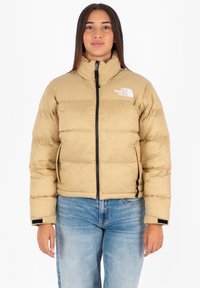 Beige pufferjack met hoge kraag, rits aan de voorkant en logo op de borst; heeft horizontale stiksels en zijzakken. Gecombineerd met blauwe jeans.