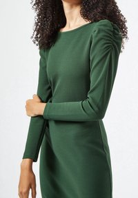 Vihreä, teksturoitu mekko pitkillä hihoilla ja pyöreällä pääntiellä. Sisältää koottua yksityiskohtaa olkapäällä lisäulottuvuuden tarjoamiseksi.