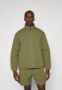 Padded olive green jacket with a high collar, full front zipper, and elastic cuffs. Worn over matching shorts. Smooth fabric texture.