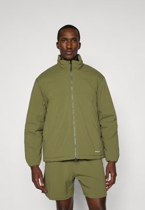 Veste olive verte rembourrée avec un col haut, une fermeture éclair sur toute la hauteur à l'avant et des poignets élastiques. Portée sur un short assorti. Texture de tissu lisse.