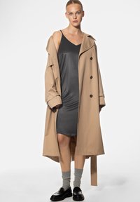 Un trench-coat beige par-dessus une robe gris foncé, complété par des chaussettes grises et des mocassins noirs. Le manteau présente des boutons et une coupe ample.