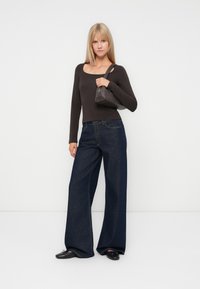 Kvinne i svart langermet topp og vide mørke jeans holder en liten svart håndveske over den ene skulderen, stående mot en enkel hvit bakgrunn.