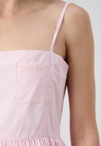 Rosa ermeløs kjole med en figursydd liv, samlet skjørt og frontlomme med brodert merkenavn. Glatt bomullsstoff.