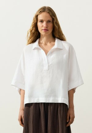 Femeie cu păr lung, ondulat și blond, purtând o cămașă albă lejeră cu guler și pantaloni largi maro cu dungi fine, pe un fundal simplu.