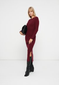 Burgunderfarget ribbestrikket kjole med båtutringning, lange ermer og sidesplitter, sammen med en svart clutch og svarte støvler til knærne.