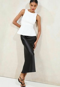 Witte peplum top met verticale naaddetails, gecombineerd met een strakke zwarte satijnen maxirok. Model draagt zwarte slippers. Effen achtergrond.