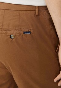 Pantaloni in cotone marrone con tasca posteriore, dotati di chiusura a bottone e di una piccola etichetta blu con la scritta "Faconnable". Texture liscia, design casual.