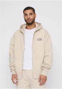 Bež dukserica na patent od mekane tkanine, s prednjim džepom i malim logotipom na lijevoj strani. Kapuljača s detaljem na vezice.