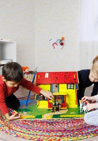 Bunte Holzhütte mit rotem Dach, gelben Wänden und grünem Sockel. Zwei Kinder spielen mit Figuren auf einem runden, strukturierten Teppich.