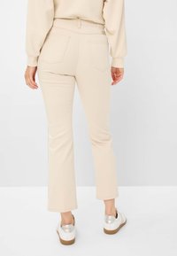 Pantaloni beige a campana con tre tasche posteriori, texture liscia e una vita. Indossati con scarpe bianche con accenti in argento.