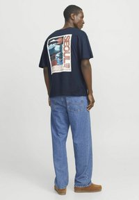Marineblå t-shirt med et farverigt grafisk design af Seoul på ryggen. Sammen med lyseblå denimjeans og brune sko.
