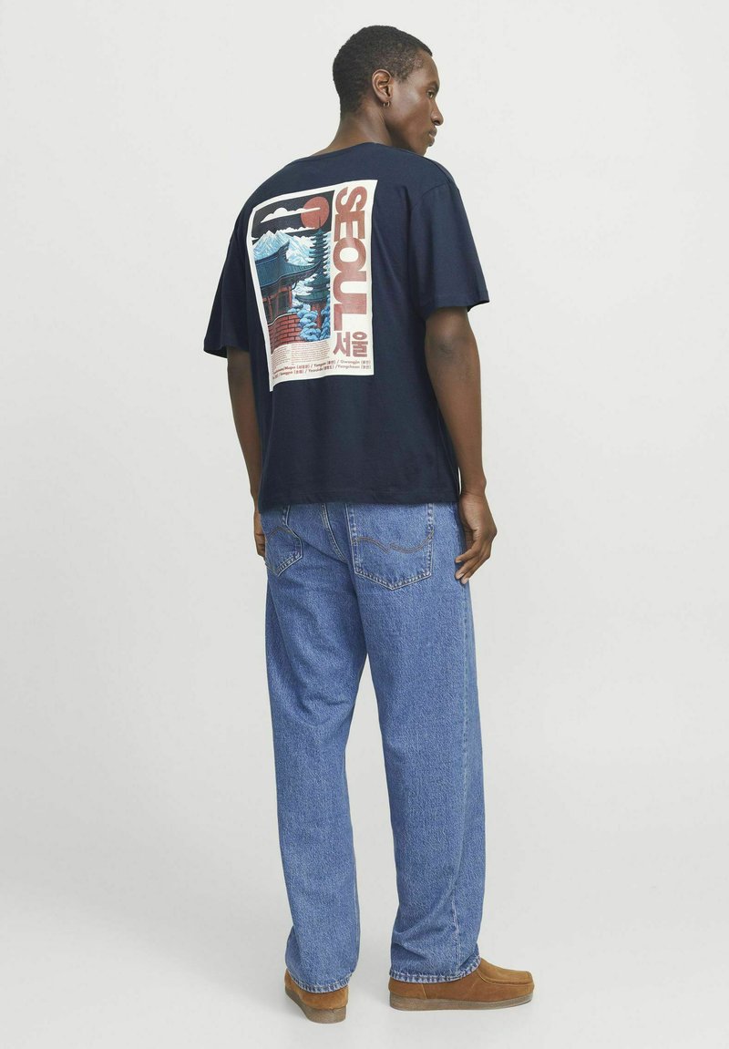Marineblå t-shirt med et farverigt grafisk design af Seoul på ryggen. Sammen med lyseblå denimjeans og brune sko.