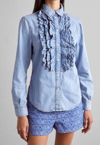 Ljusblå denimskjorta med volangdetaljer fram, knäppning med knappar och långa ärmar. Matchas med blåa shorts med ett upprepat logomönster.