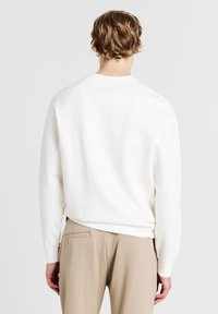 Felpa bianca con maniche lunghe, collo rotondo, texture liscia. Indossata con pantaloni beige, che mostrano una vestibilità rilassata e un design minimalista.