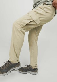 Beige Cargo-Hosen aus Baumwollmischung. Verfügen über mehrere Seitentaschen, geraden Schnitt und leichtes Material. Kombiniert mit grauen Sportschuhen.